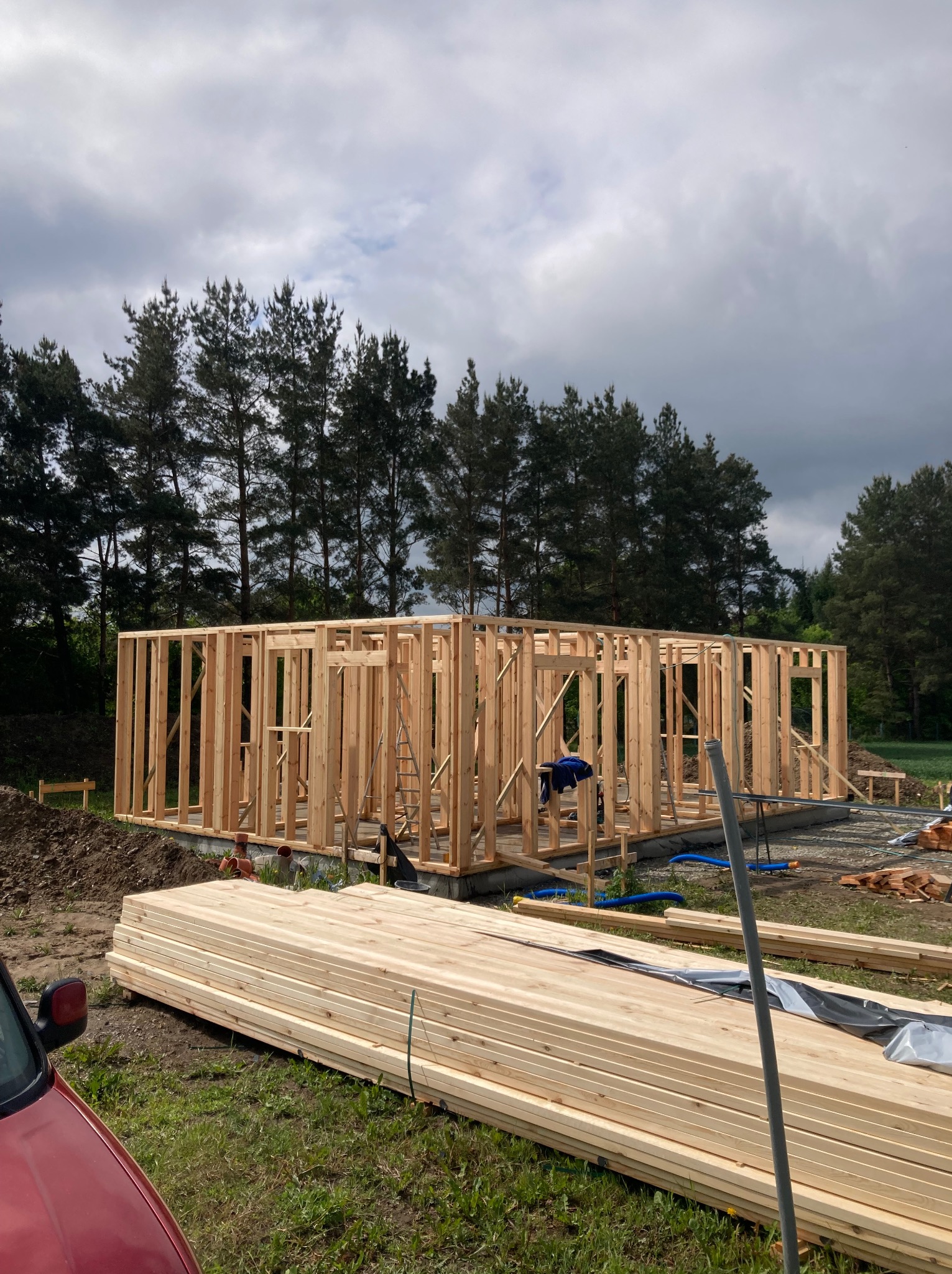 Wznoszenie konstrukcji domu szkieletowego: drewniany szkielet na fundamencie, stos desek w pierwszym planie, las w tle pod pochmurnym niebem.