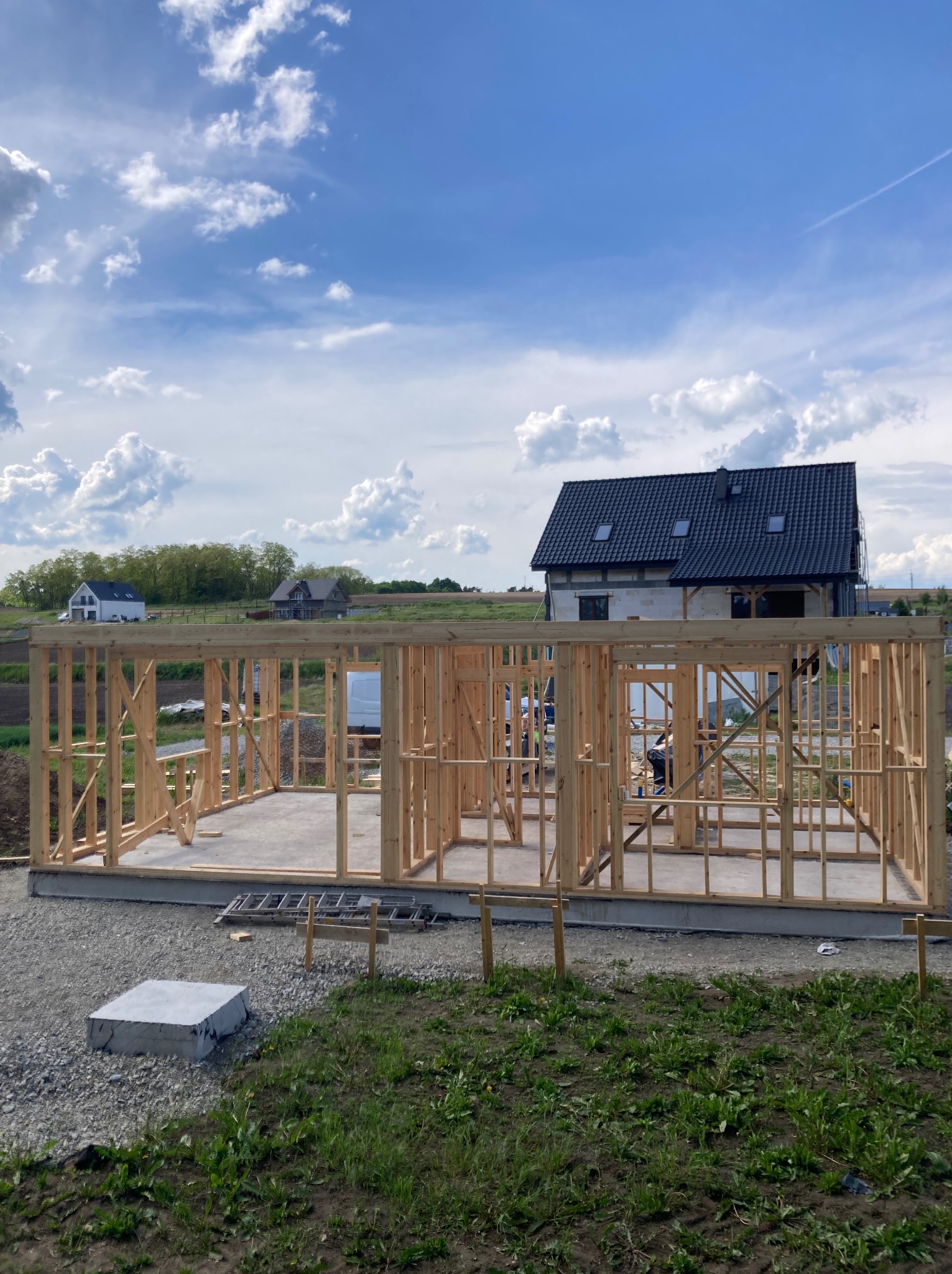 Wznoszenie szkieletu domu jednorodzinnego na fundamencie. Widoczne drewniane elementy konstrukcyjne i częściowo wykończony dom w tle. Letnie, słoneczne warunki.