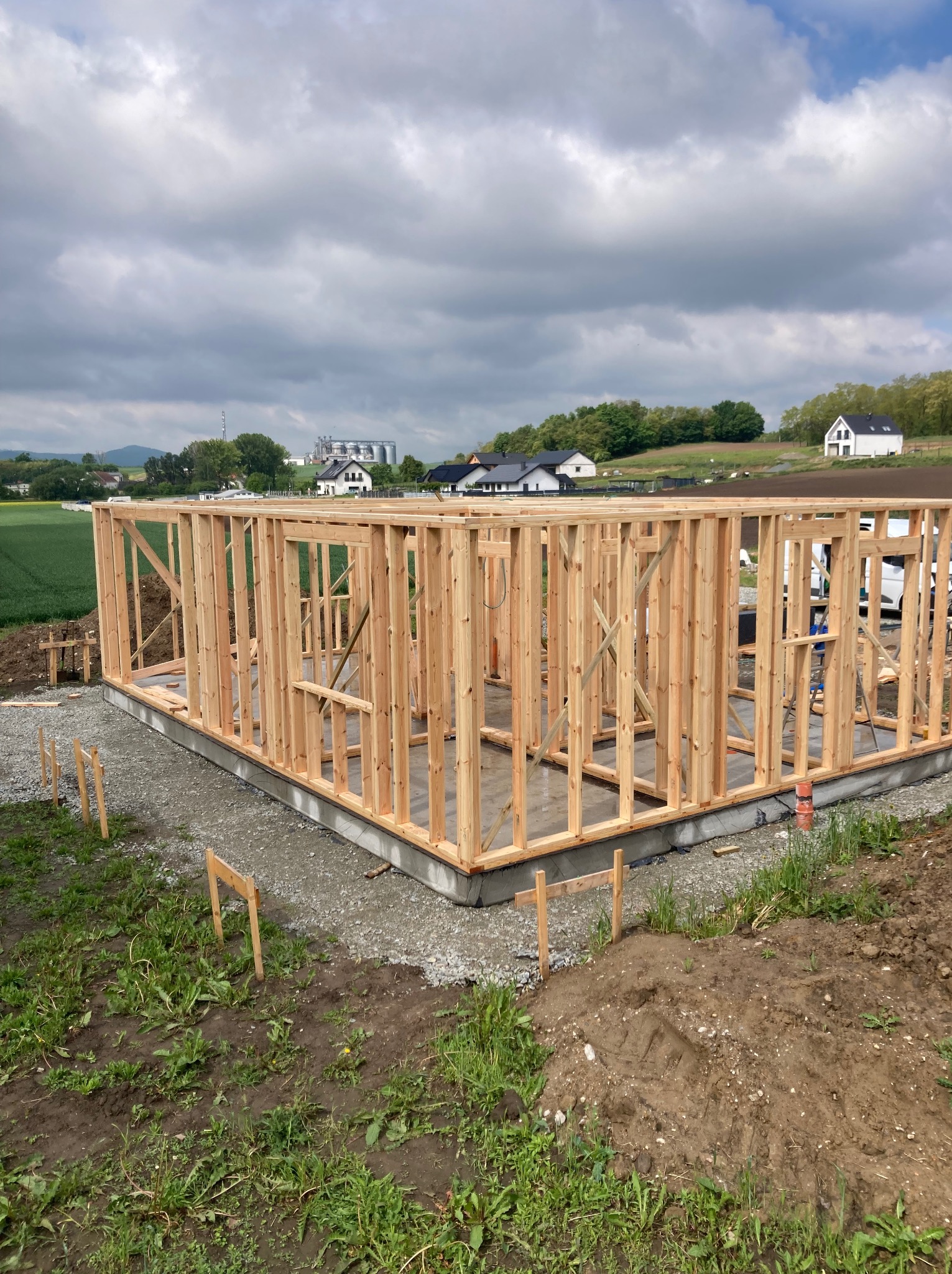 Konstrukcja szkieletowa domu w trakcie budowy, widok z zewnątrz. Fundament betonowy, drewniane ściany, tło: zielone pole, budynki mieszkalne i pochmurne niebo.