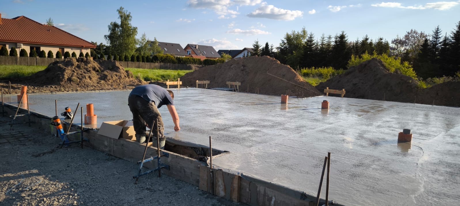 Wylewka fundamentowa pod budowę, pracownik wygładza powierzchnię betonu. Widoczne szalunki, wystające rury i przygotowany teren pod budowę domu.