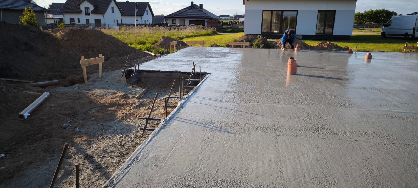 Wylany fundament pod dom szkieletowy. Widoczne wystające pręty zbrojeniowe, fragment budynku w tle i pracownik wygładzający powierzchnię betonu.