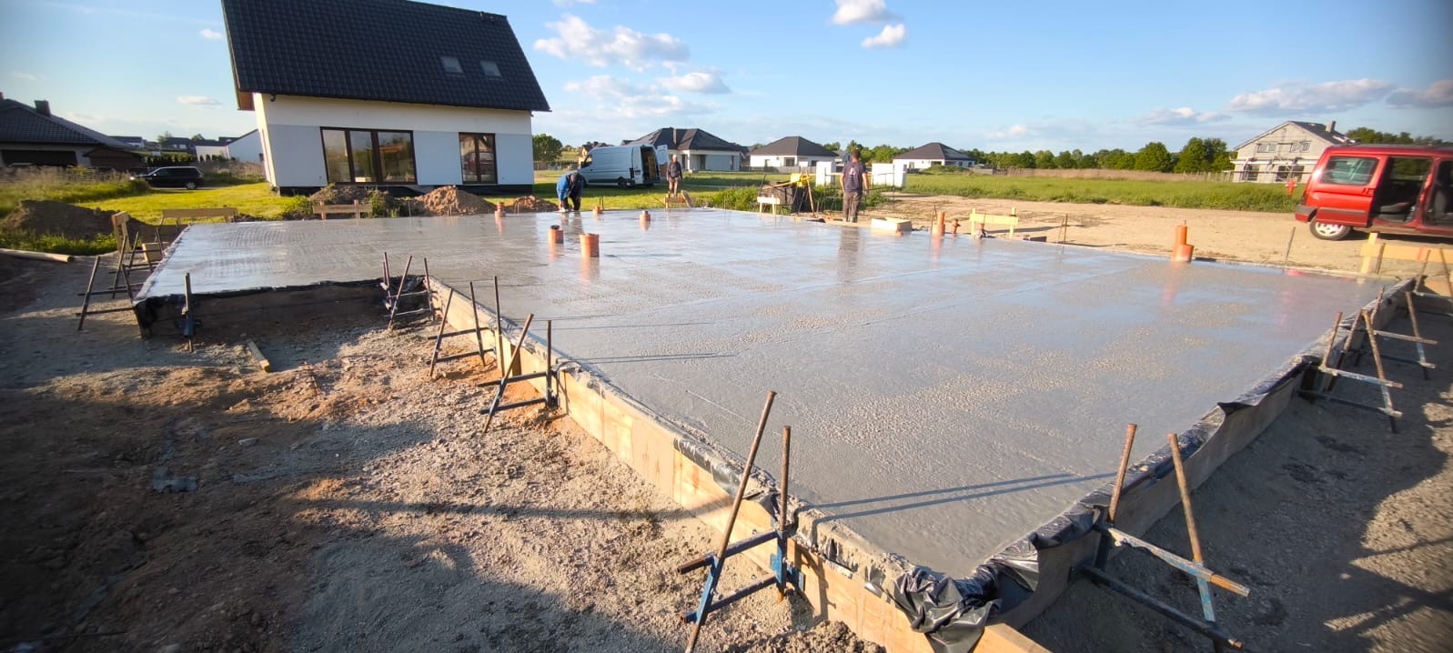 Wylany fundament pod dom szkieletowy na działce z widokiem na inne domy. Widoczne szalunki, zbrojenie i świeży beton. W tle pracownicy budowlani.