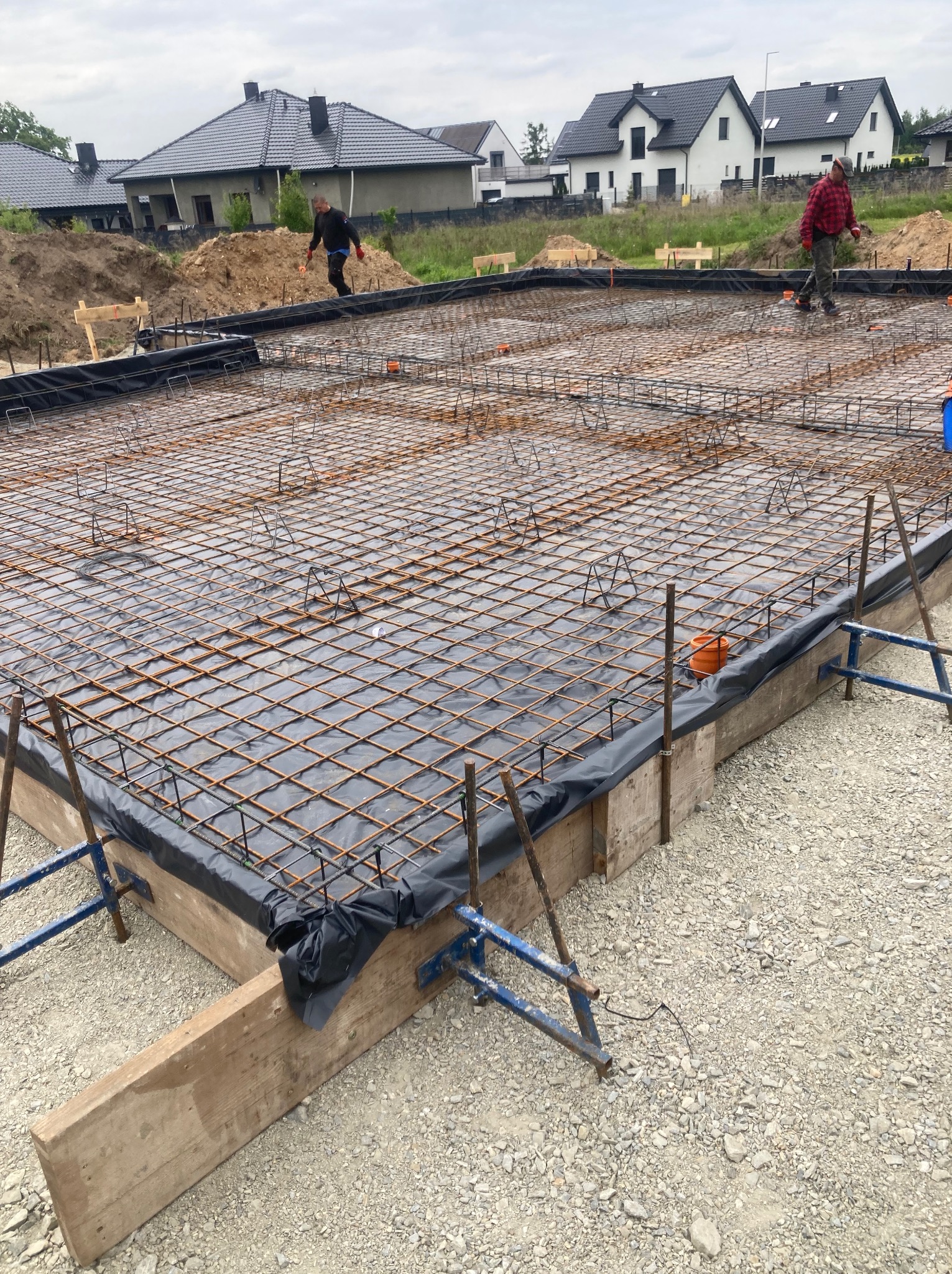 Zbrojenie fundamentu pod budowę domu szkieletowego, widoczne szalunki i folia izolacyjna. Dwóch pracowników na budowie, w tle domy jednorodzinne.