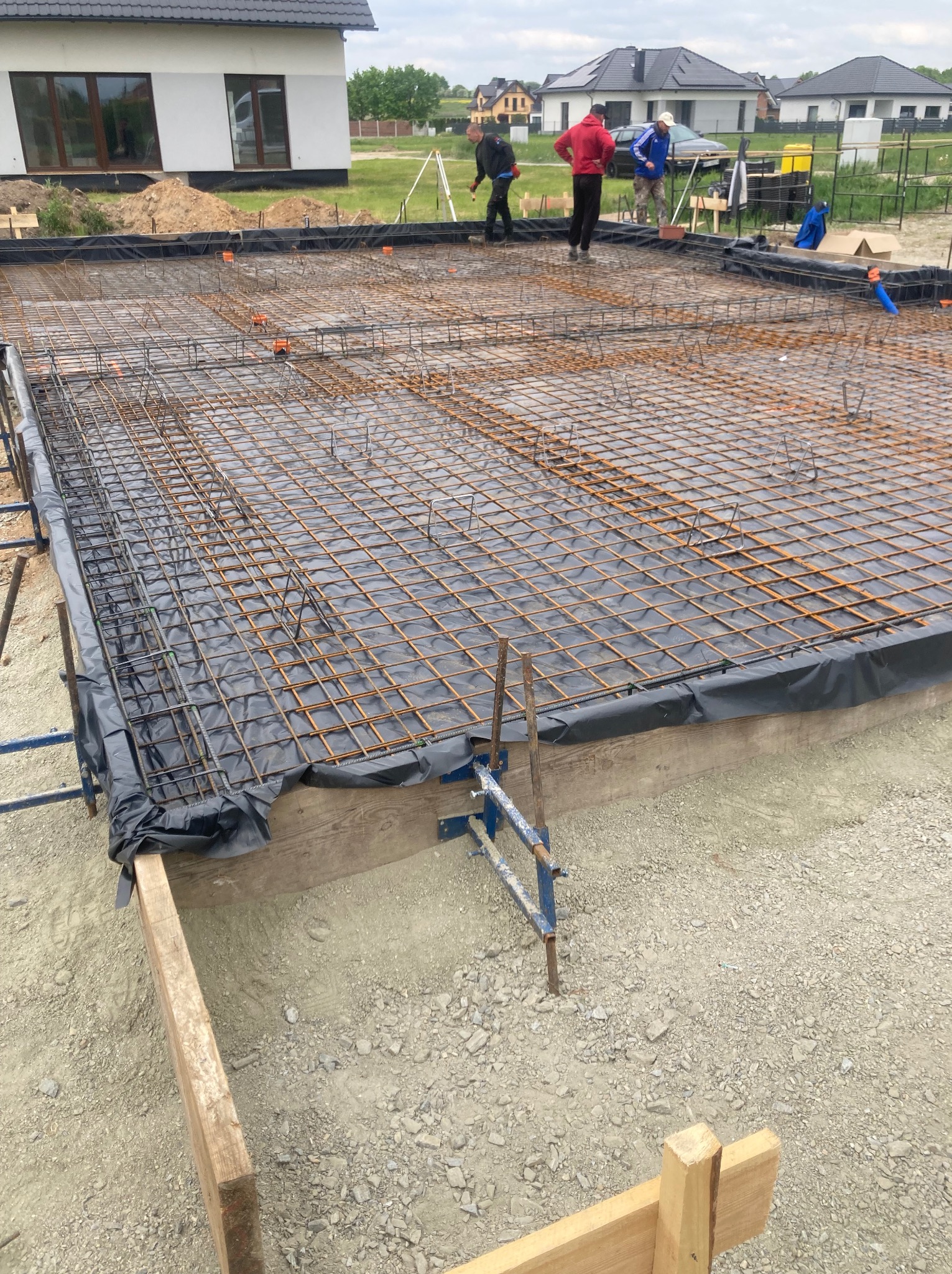 Zbrojenie fundamentów pod dom szkieletowy, zabezpieczone folią. Widoczni pracownicy budowlani w tle, dom w stanie surowym zamkniętym.