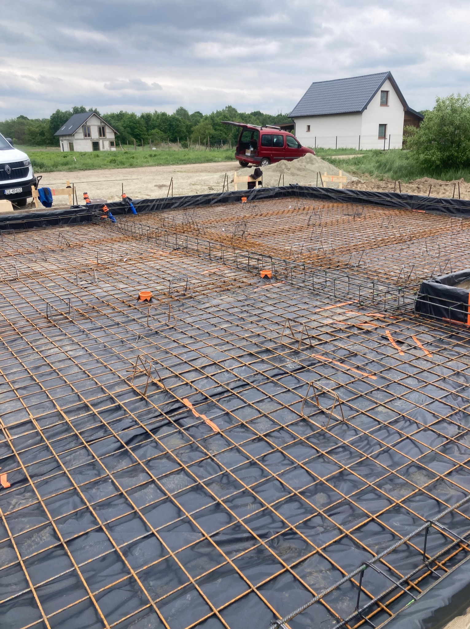 Siatka zbrojeniowa na czarnej folii, przygotowana do wylewki fundamentów pod budowę domu. W tle domy w różnym stadium budowy i czerwony samochód.