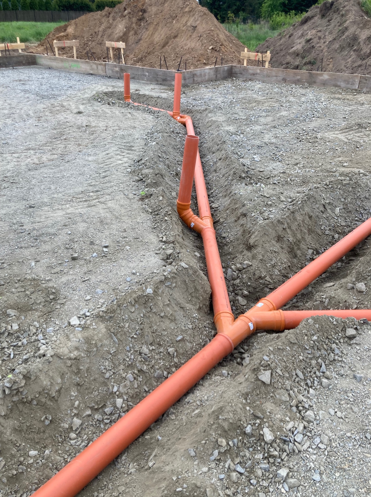 Pomarańczowe rury kanalizacyjne w wykopie na fundamencie pod budowę. Widoczne złącza typu Y. W tle szalunek i ziemia.
