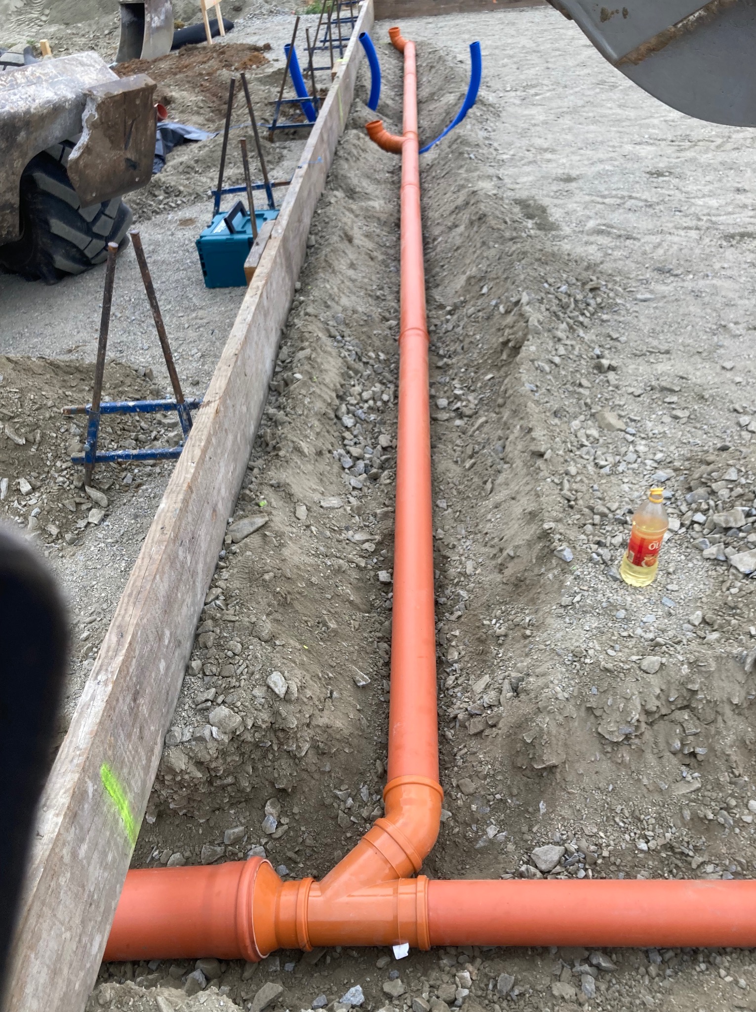 Pomarańczowe rury kanalizacyjne w wykopie, przygotowane do zasypania. Widoczne drewniane szalunki, fragment koparki i butelka oleju. Prace ziemne.