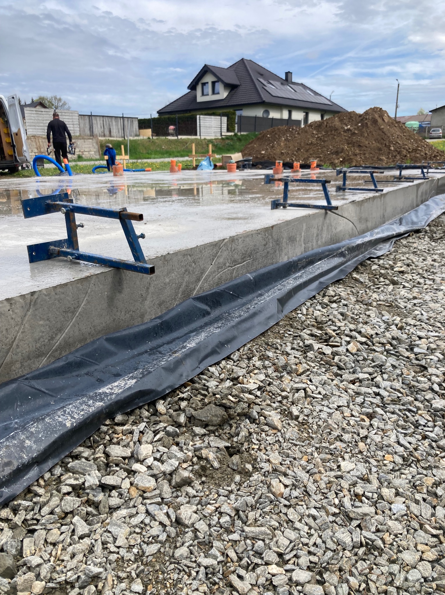 Betonowa podstawa pod budowę domu z widocznymi elementami konstrukcyjnymi i folią hydroizolacyjną. W tle dom z ciemnym dachem i stertą ziemi.