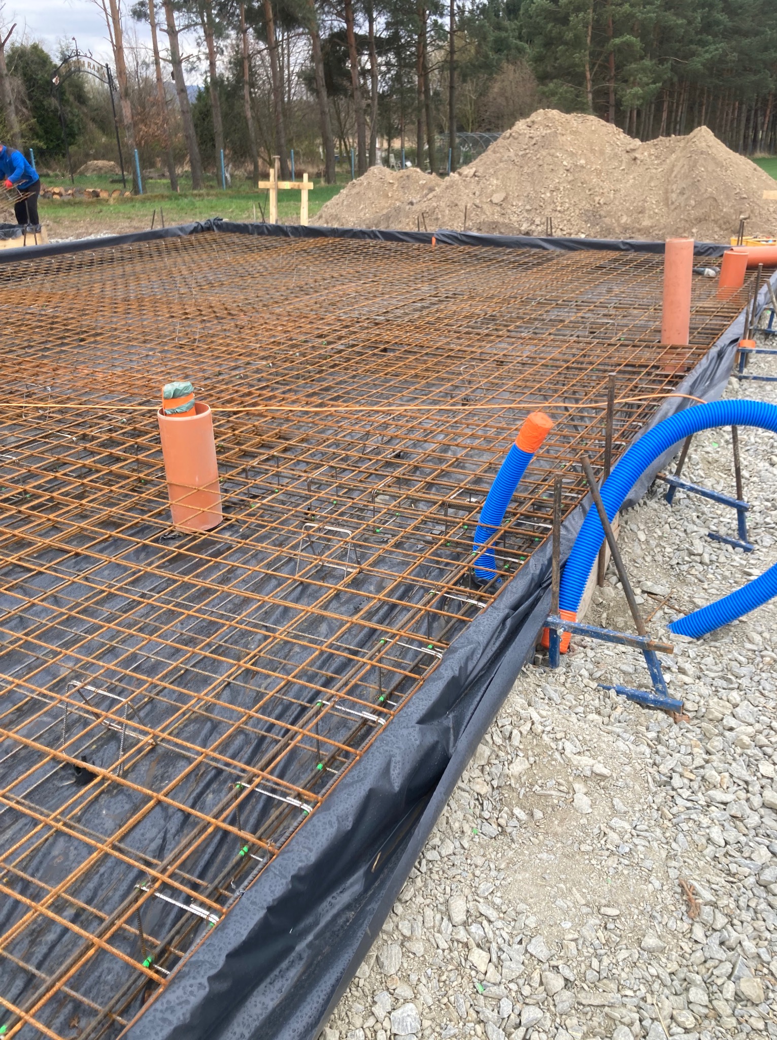 Zbrojenie fundamentów pod budowę domu szkieletowego z widocznymi rurami instalacyjnymi i podsypką z kamienia. Prace przygotowawcze w toku.