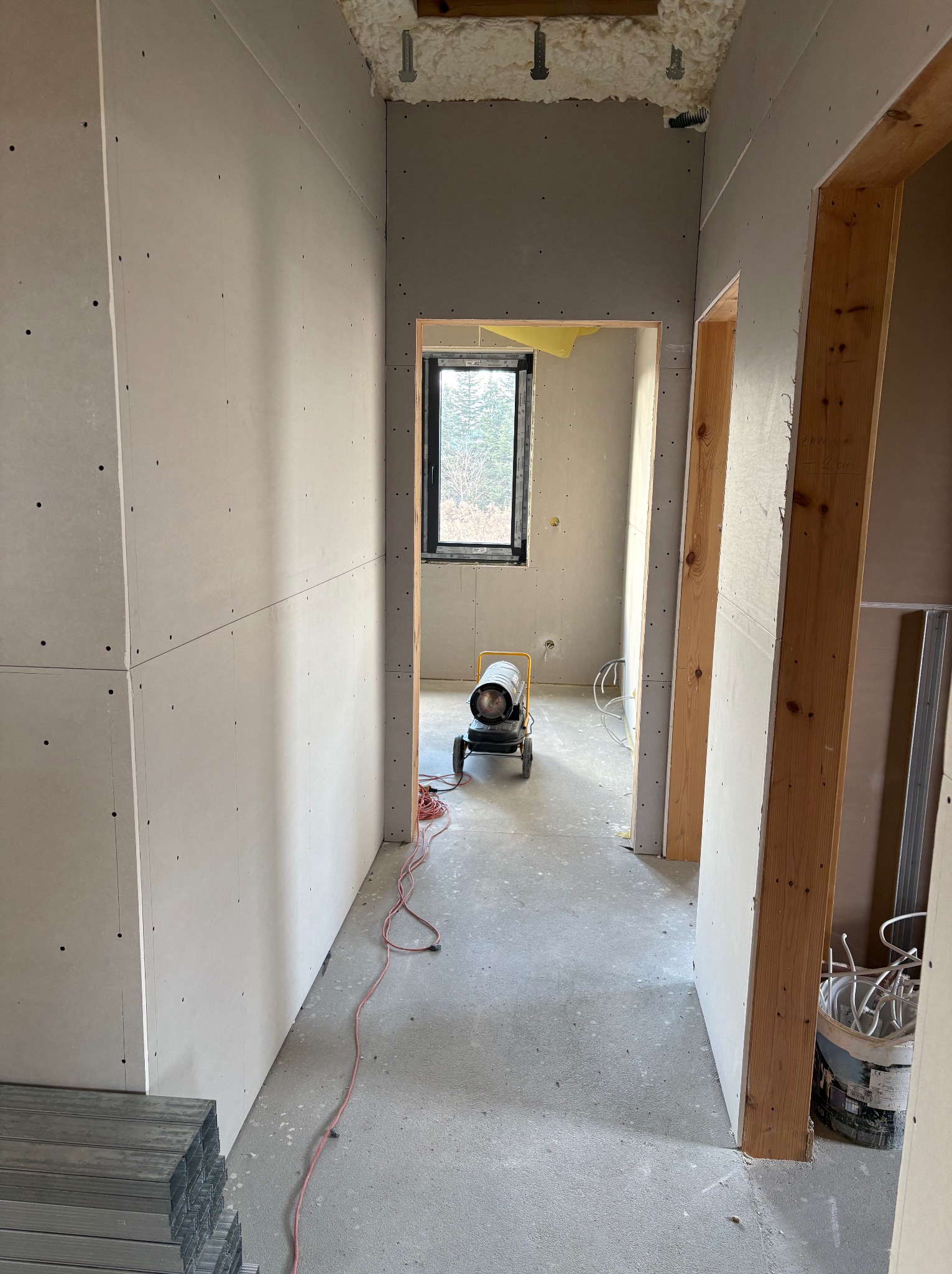 Wnętrze w trakcie budowy domu szkieletowego: ściany z płyt gipsowo-kartonowych, widoczne drewniane ramy drzwi, okno i nagrzewnica. Etap wykończeniowy.