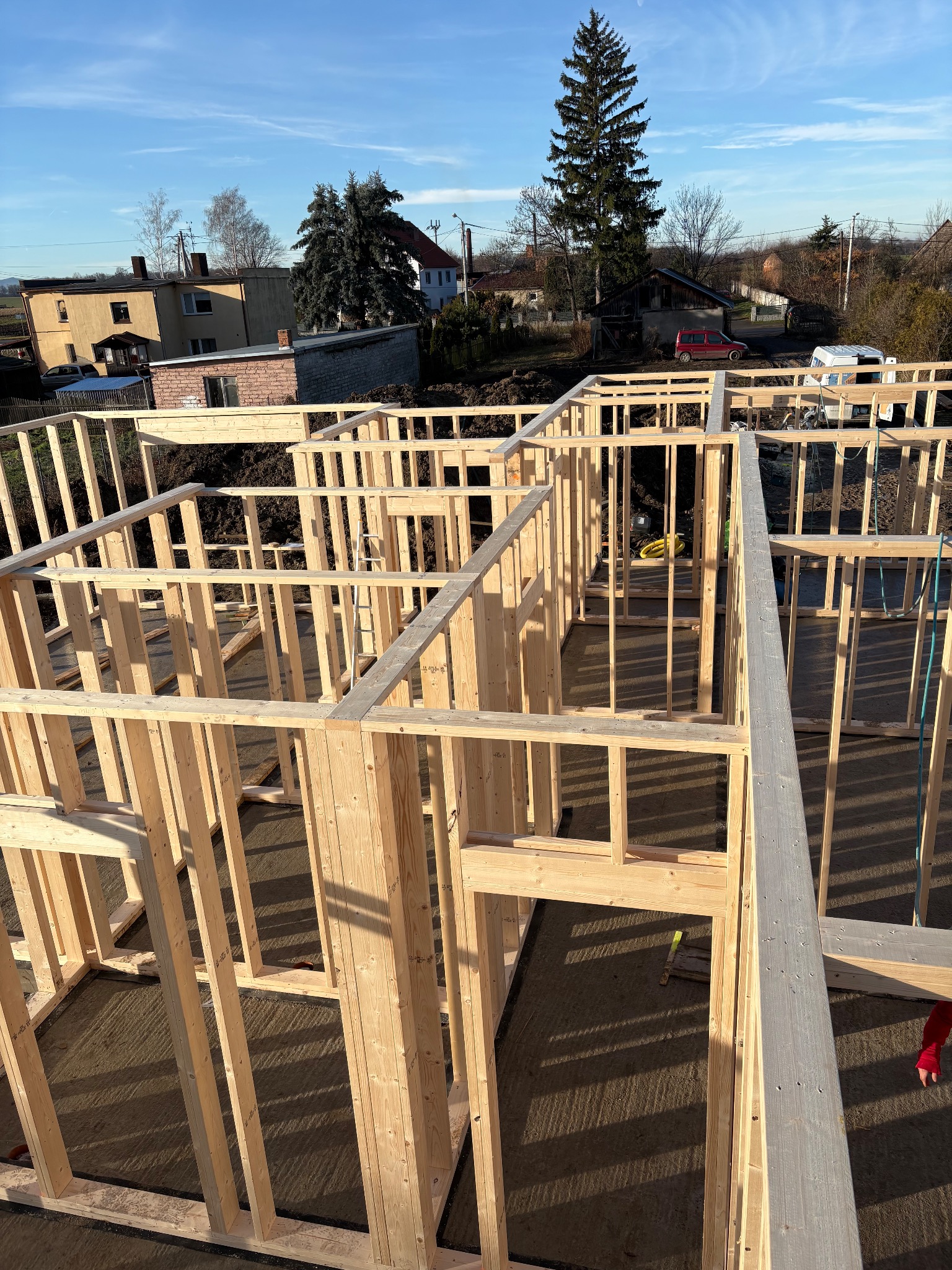 Wysoki kąt na drewniany szkielet domu w budowie, z widocznymi belkami i słupami. W tle zabudowa jednorodzinna i drzewa pod błękitnym niebem.
