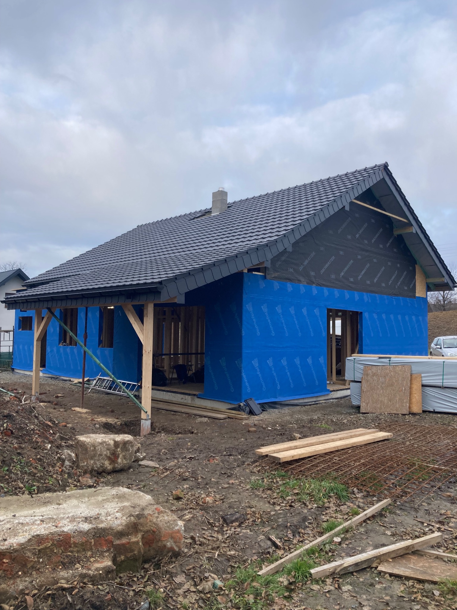 Budowa domu szkieletowego w trakcie realizacji: widoczne drewniane elementy konstrukcyjne, niebieska membrana i czarna dachówka. Częściowo wykończony dach i ściany.