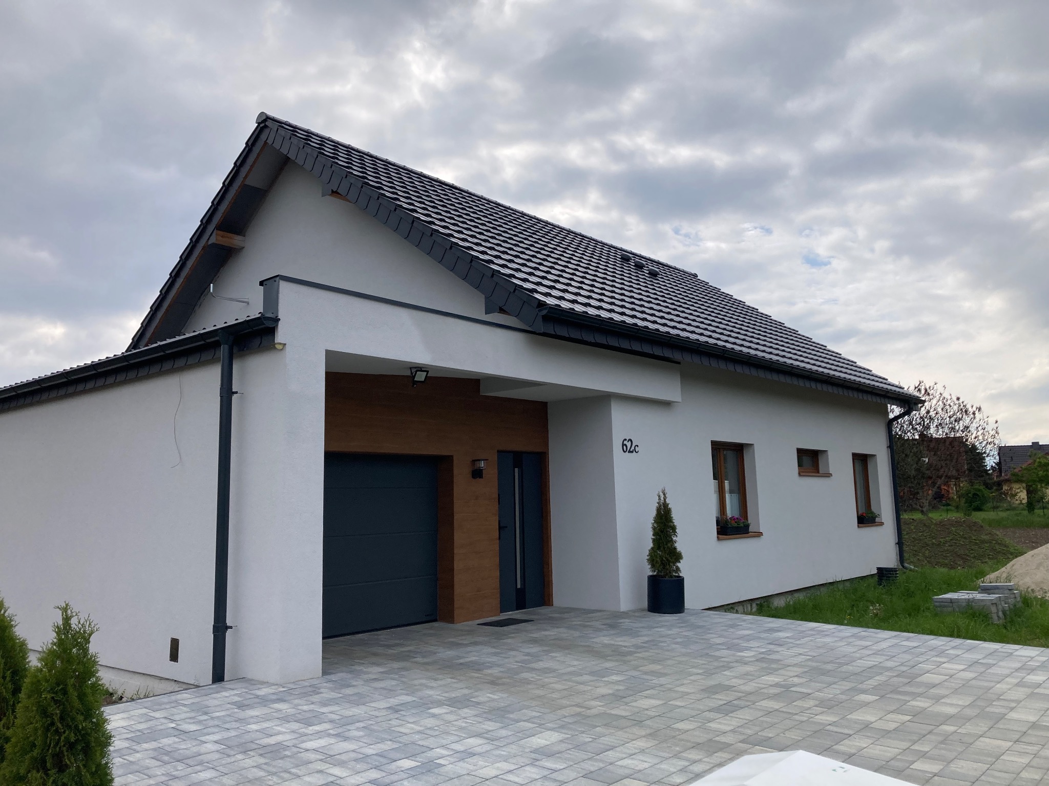 Nowoczesny dom z garażem i drewnianymi akcentami, wykończony jasną elewacją i ciemną dachówką. Podjazd wyłożony kostką brukową, małe drzewko w donicy przy wejściu.