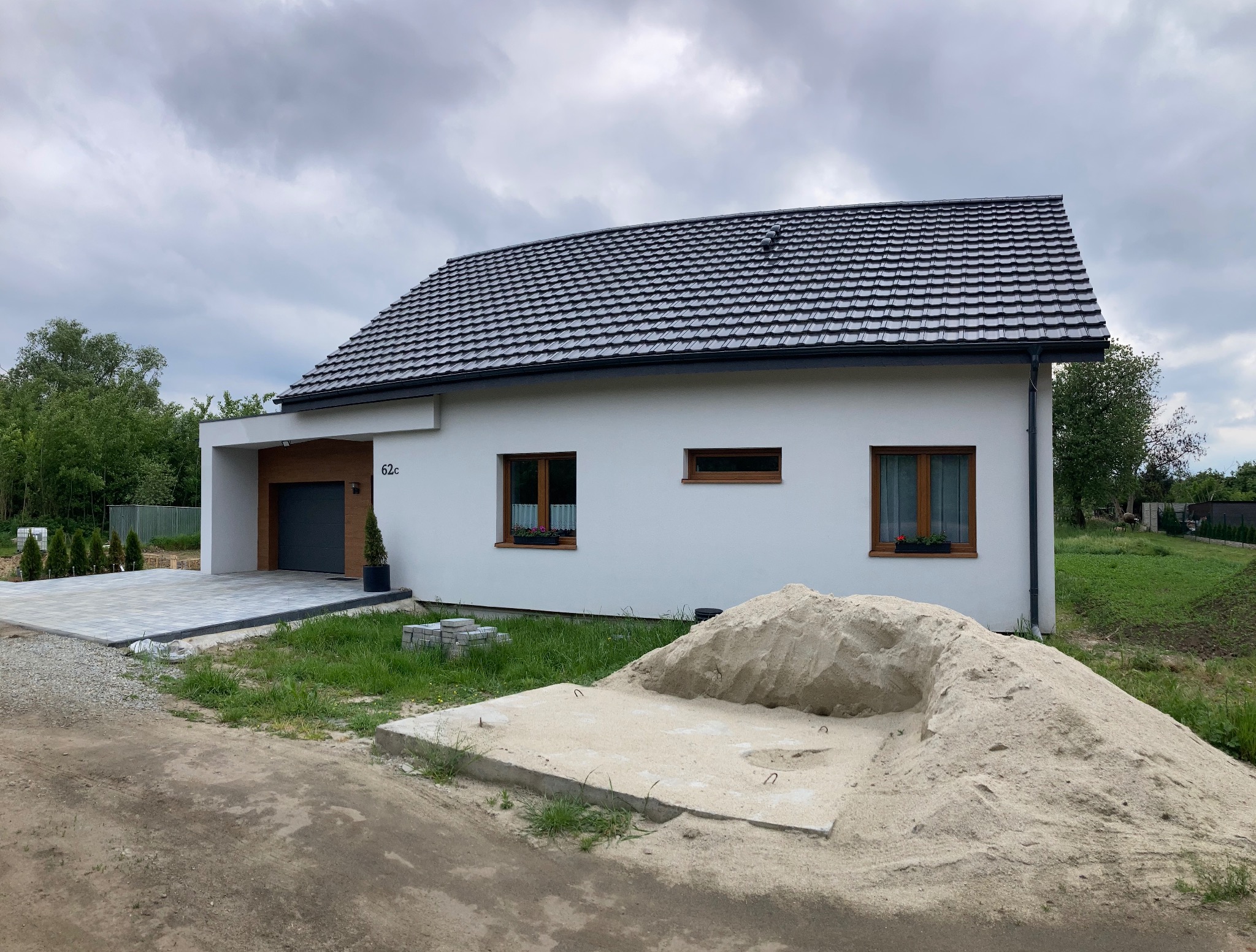 Nowoczesny, parterowy dom z białymi ścianami i grafitową dachówką. Widoczny podjazd z kostki brukowej i przygotowany piasek budowlany. Numer domu 62c.