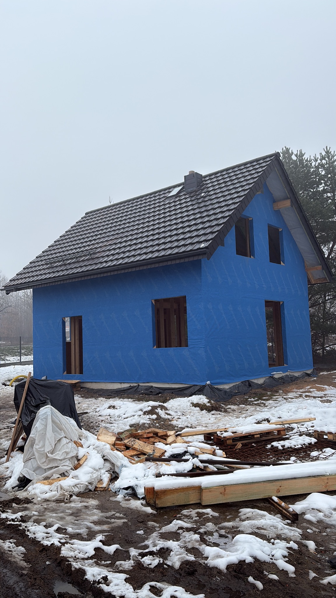 Niedokończony dom z niebieską membraną i ciemnym dachem w zimowej scenerii. Widoczne drewniane elementy konstrukcyjne i resztki materiałów budowlanych.