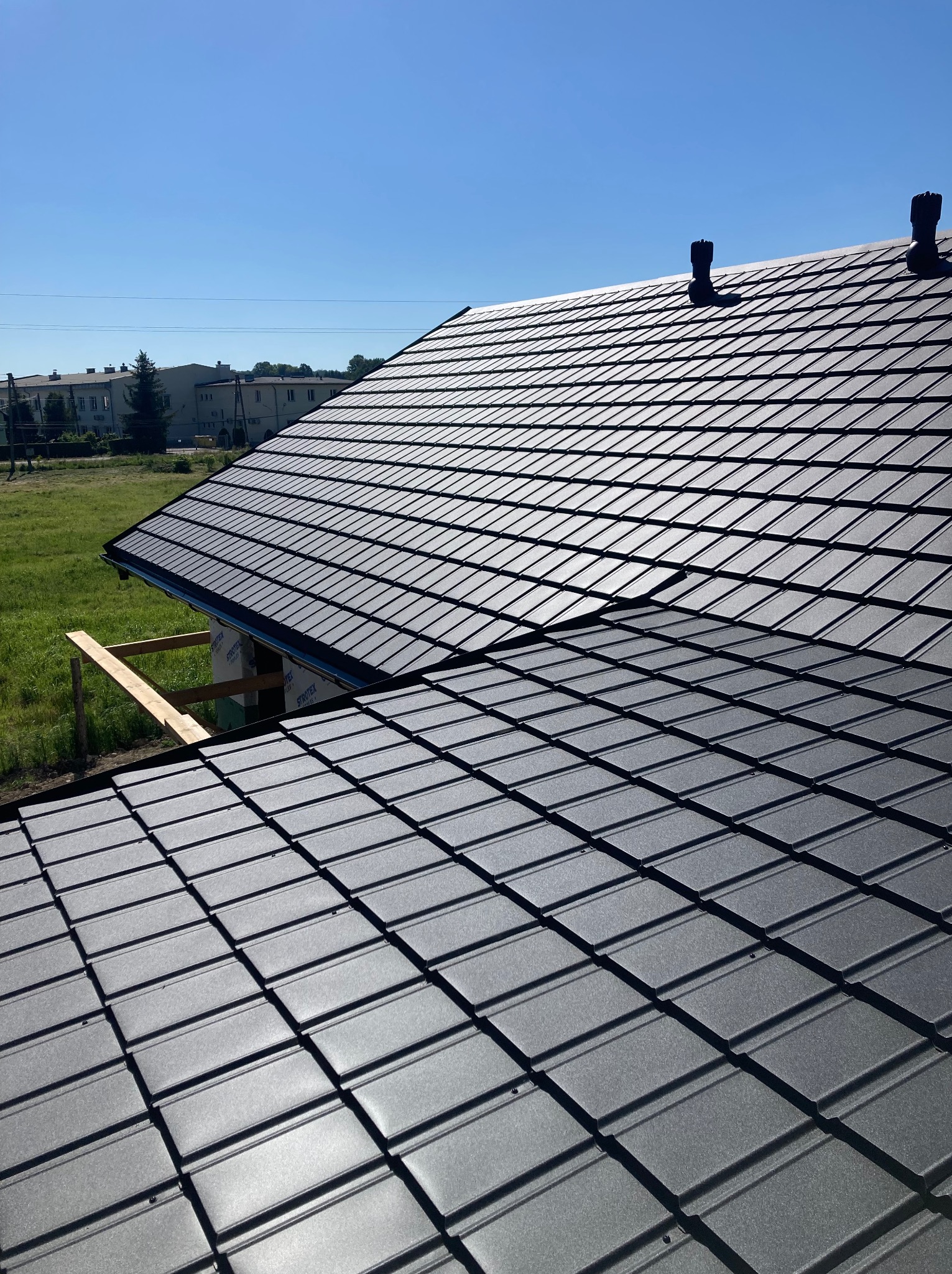 Ciemnoszara dachówka ceramiczna na ukończonym dachu domu szkieletowego w słoneczny dzień. Widoczne kominy wentylacyjne i fragment konstrukcji.