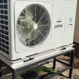 Biała jednostka zewnętrzna pompy ciepła Galmet Prima, zamontowana na czarnym, metalowym stelażu przed budynkiem, widoczny wentylator i oznaczenie R32.