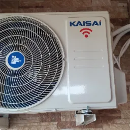 Zewnętrzna jednostka klimatyzacji marki Kaisai zamontowana na brązowo-beżowej ścianie z imitacji kamienia, widoczne białe rury i uchwyty montażowe.