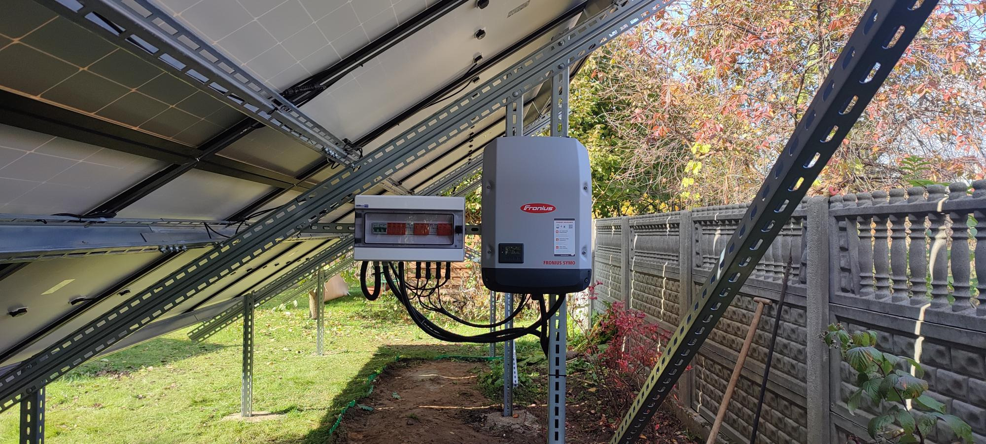 Instalacja paneli fotowoltaicznych z widocznym inwerterem Fronius i skrzynką elektryczną na tle jesiennego ogrodu z betonowym ogrodzeniem.