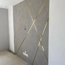 Dekoracyjna ściana z szarego tynku strukturalnego z geometrycznym wzorem z mosiężnych listew, widoczne gniazdka elektryczne.