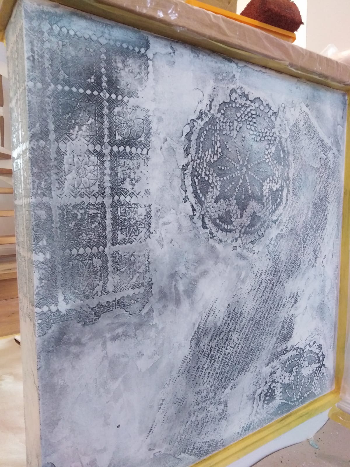 Dekoracyjna ściana z efektem przetarć i wzorami imitującymi koronki, zabezpieczona folią malarską i taśmą, w trakcie malowania.