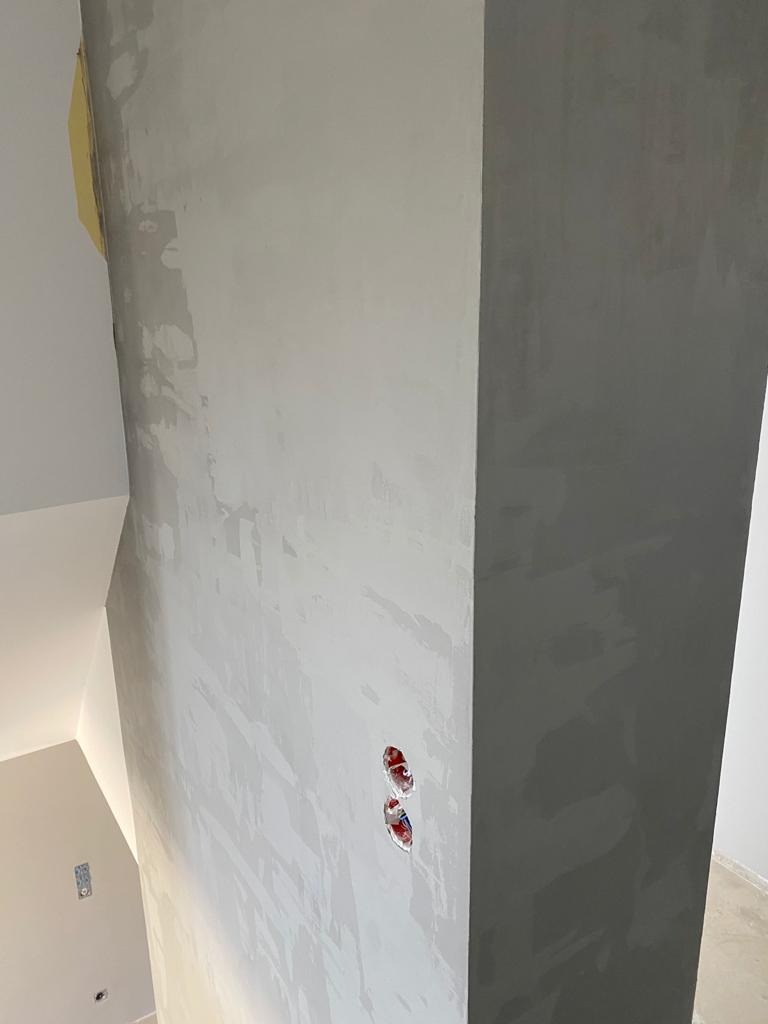 Ściana po nałożeniu gładzi szpachlowej, widoczne puszki elektryczne bez osprzętu, fragment taśmy malarskiej na górze ściany.