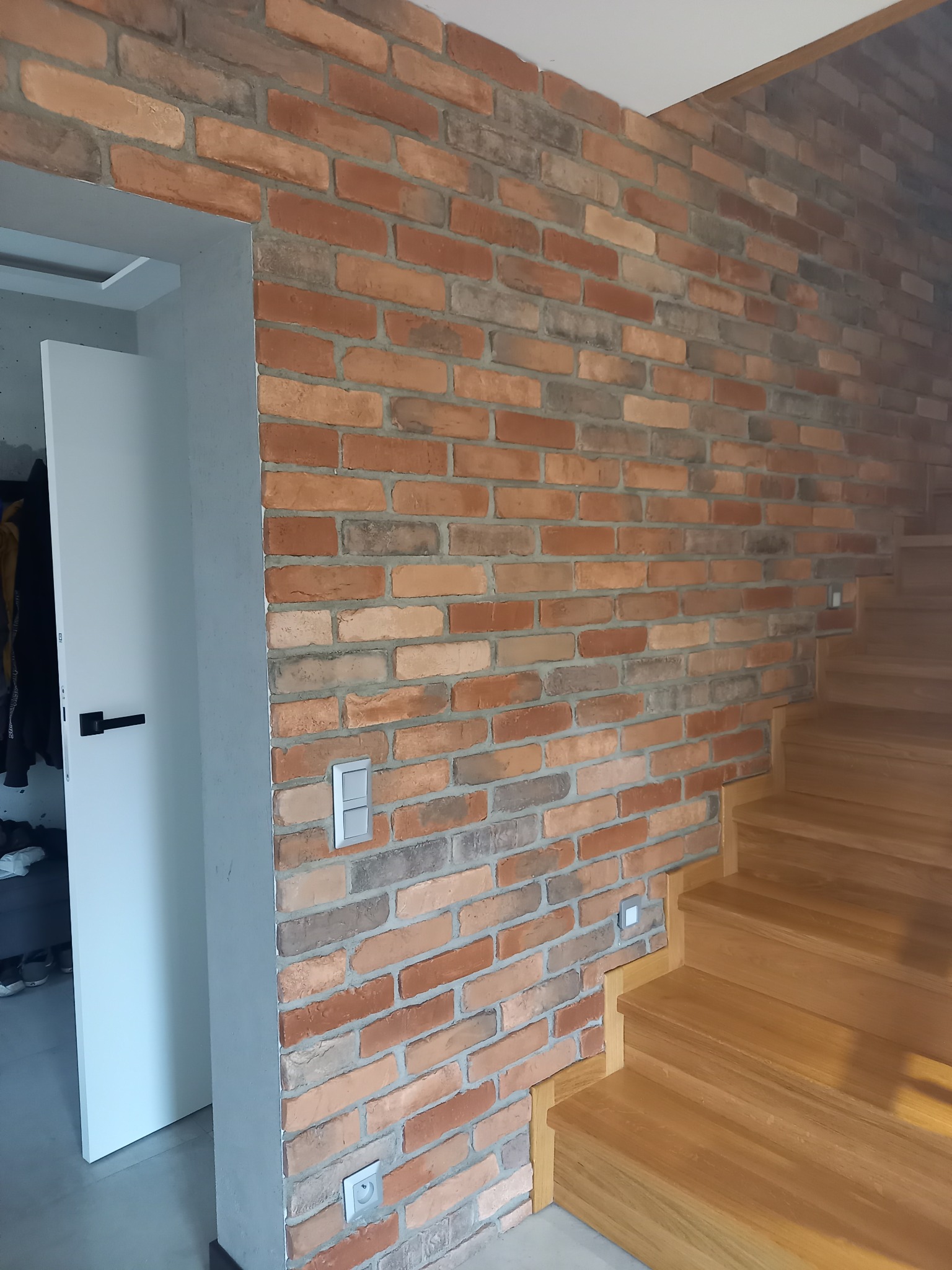 Ściana z cegły klinkierowej w odcieniach czerwieni i brązu, z widocznymi drewnianymi schodami i otwartymi drzwiami do garderoby z białymi frontami, z szarą ościeżnicą i czarną klamką.