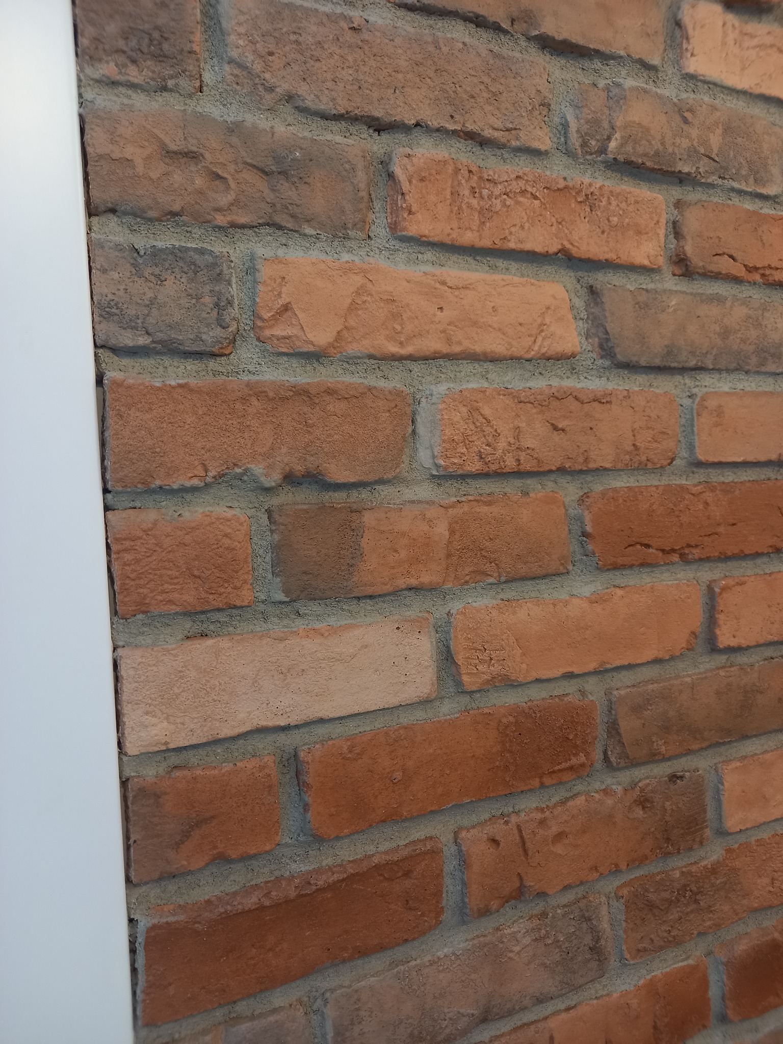 Dekoracyjna ściana z cegły klinkierowej w odcieniach czerwieni i brązu, z szarą fugą, przylegająca do białej ściany.