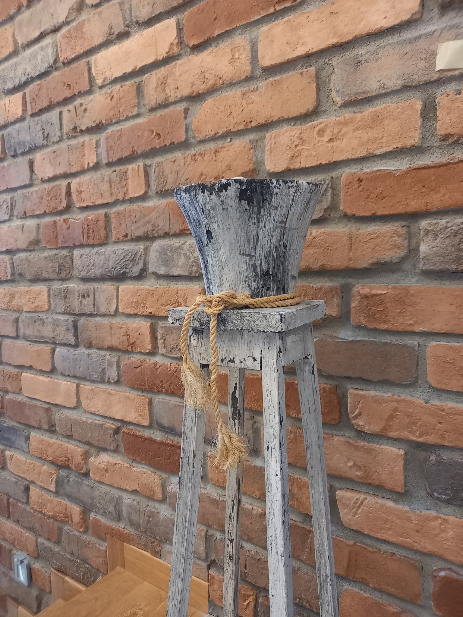 Dekoracyjna, postarzana donica w odcieniach szarości, umieszczona na drewnianym, trójnożnym postumencie, z grubym sznurem jutowym, na tle ściany z cegły klinkierowej w odcieniach czerwieni i brązu.