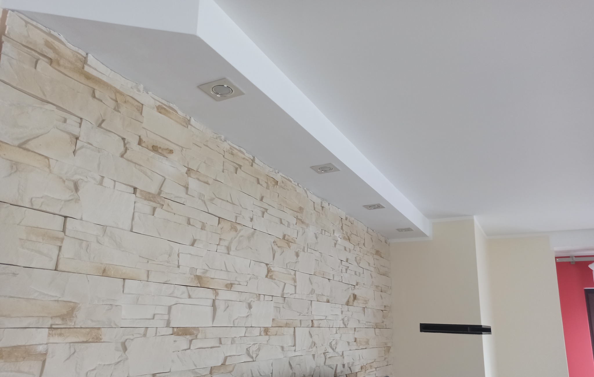 Wykończona ściana z dekoracyjnej cegły w odcieniach bieli i beżu, oświetlona trzema punktowymi lampami w suficie podwieszanym z białymi listwami.