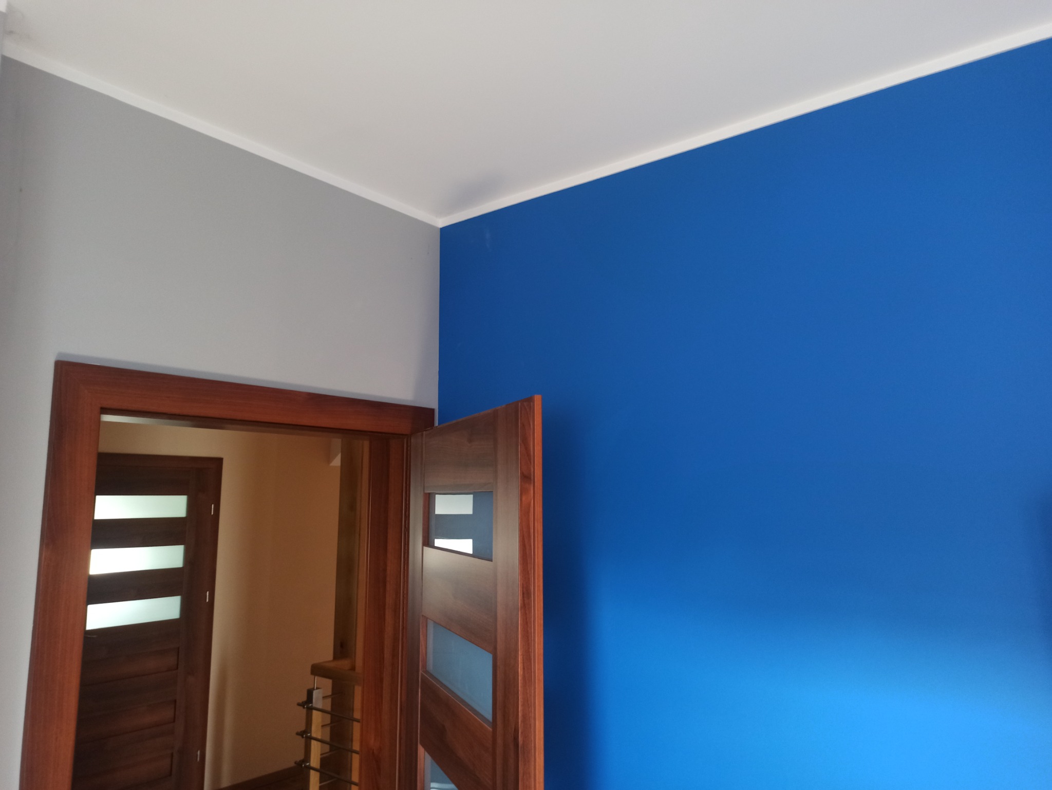 Świeżo pomalowane wnętrze z drewnianą ościeżnicą i częściowo otwartymi drzwiami. Jedna ściana w kolorze niebieskim, druga szara. Widoczny biały sufit z listwą.