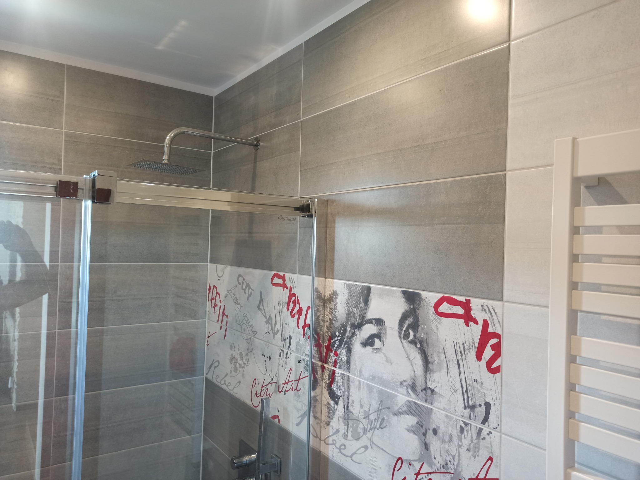 Nowoczesna łazienka z szarymi płytkami imitującymi kamień, kabiną prysznicową ze szklanymi drzwiami i deszczownicą oraz dekoracyjnymi płytkami z motywem graffiti i wizerunkiem kobiety.