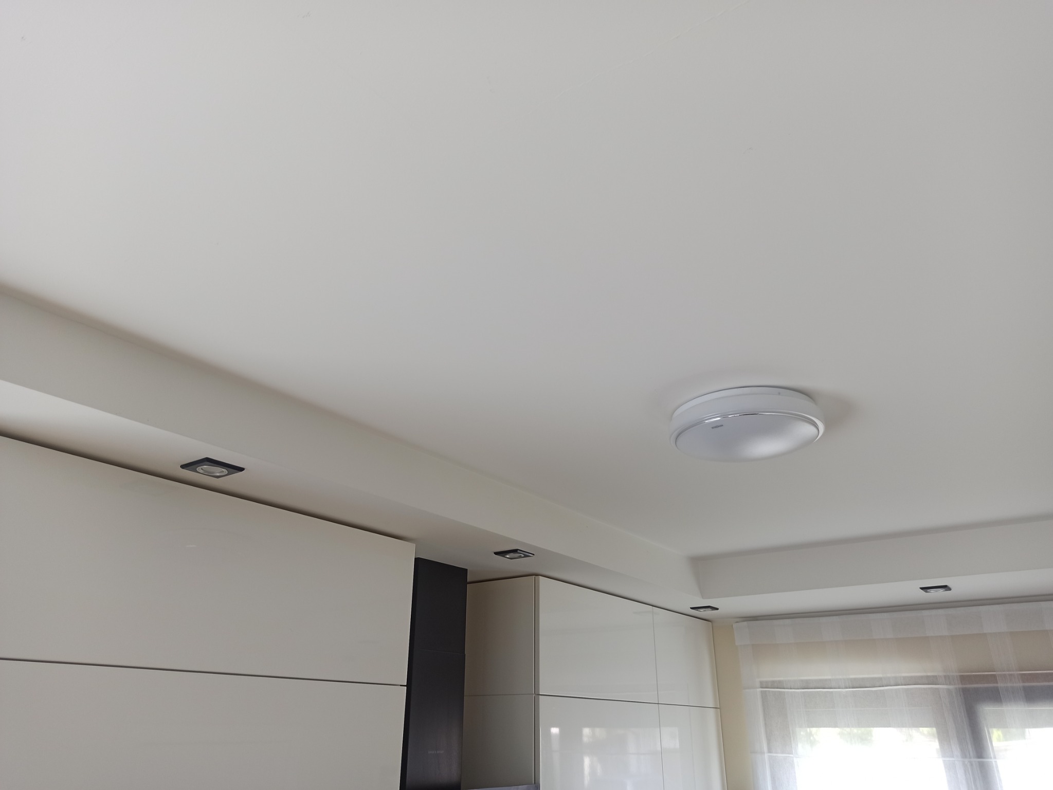 Biały sufit z zamontowanymi punktowymi lampkami halogenowymi i okrągłą lampą sufitową, widoczne fragmenty białych szafek kuchennych i okno z zasłoną.