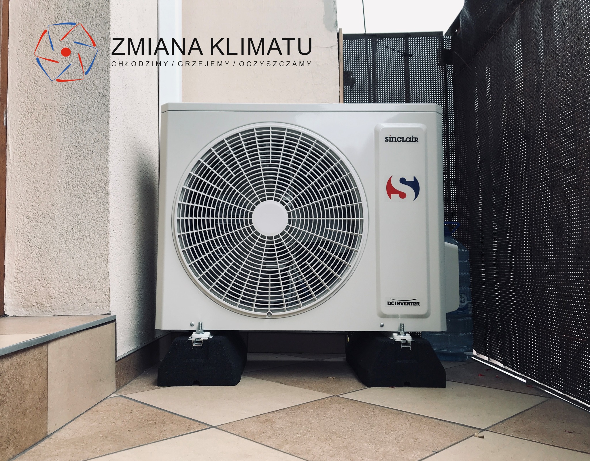 Zewnętrzna jednostka klimatyzacji Sinclair na czarnych podkładkach antywibracyjnych, umieszczona na brązowej podłodze z płytek, widoczny fragment ściany i logo firmy Zmiana Klimatu