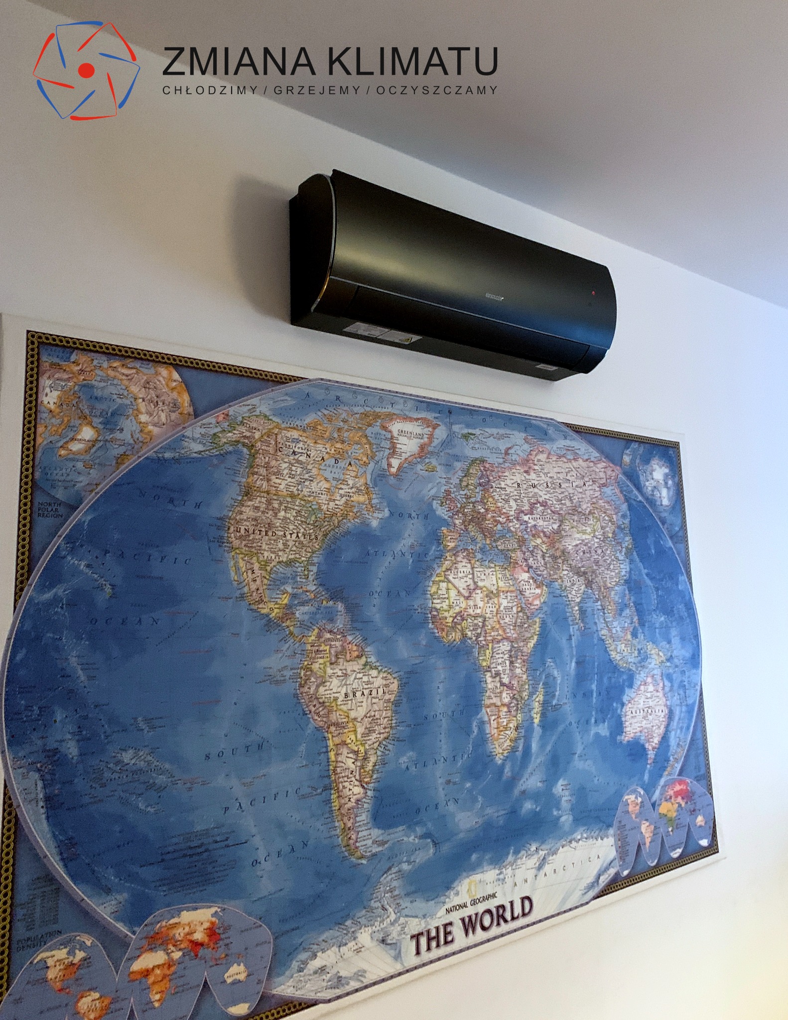 Czarny klimatyzator ścienny zamontowany nad dużą mapą świata w ramie, widoczne logo firmy na ścianie.