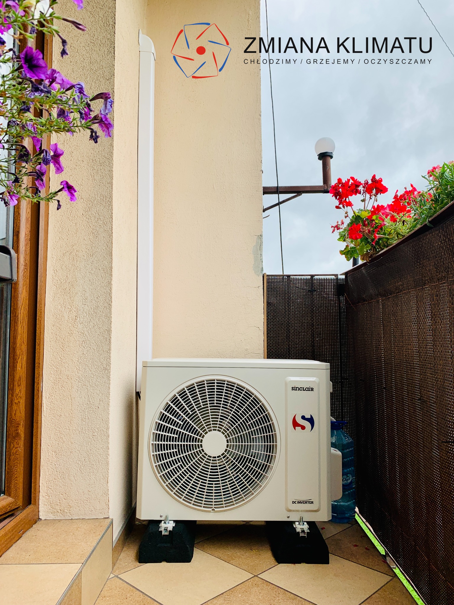 Zamontowana jednostka zewnętrzna klimatyzacji Sinclair na balkonie z kwiatami w doniczkach, widoczny odpływ skroplin.