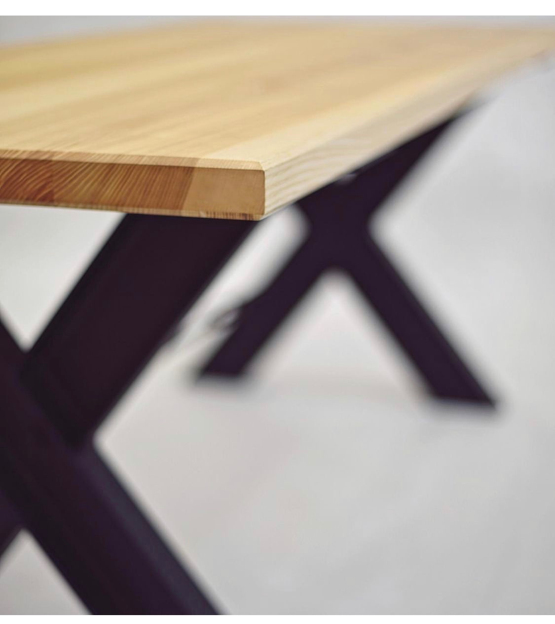 Fragment drewnianego blatu stołu z widocznym usłojeniem, wspartego na metalowej podstawie w kształcie litery X, całość na rozmytym, jasnym tle.