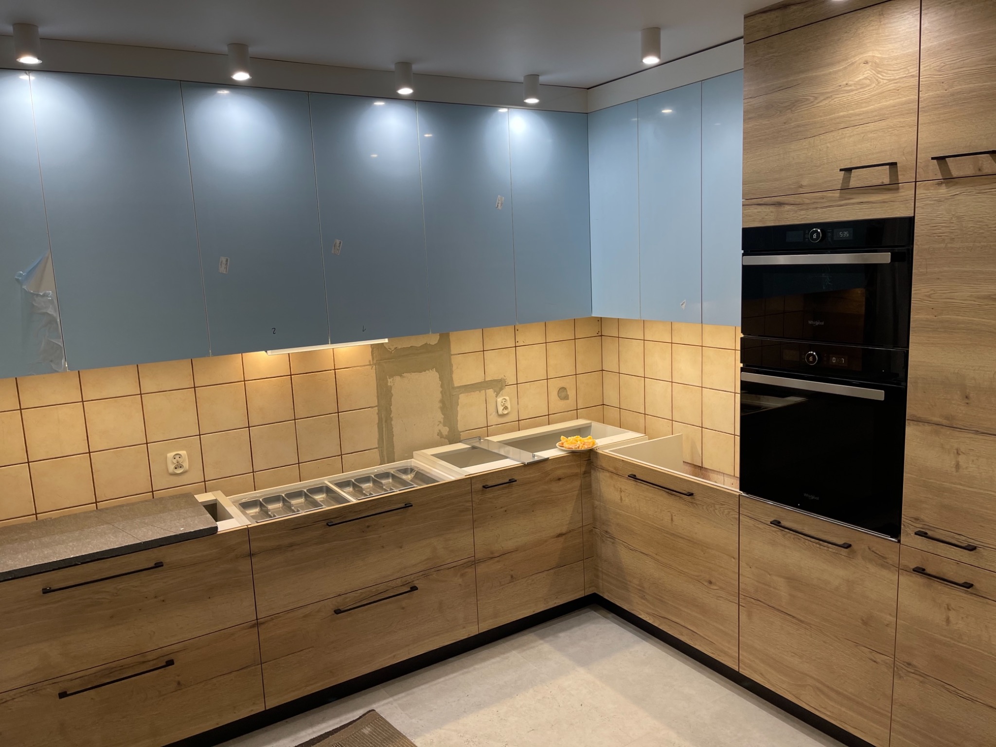 Wnętrze kuchni w trakcie montażu z niebieskimi górnymi szafkami i drewnopodobnymi dolnymi szafkami, widoczne niedokończone wykończenie ściany nad blatem z beżowych płytek oraz wbudowany piekarnik.