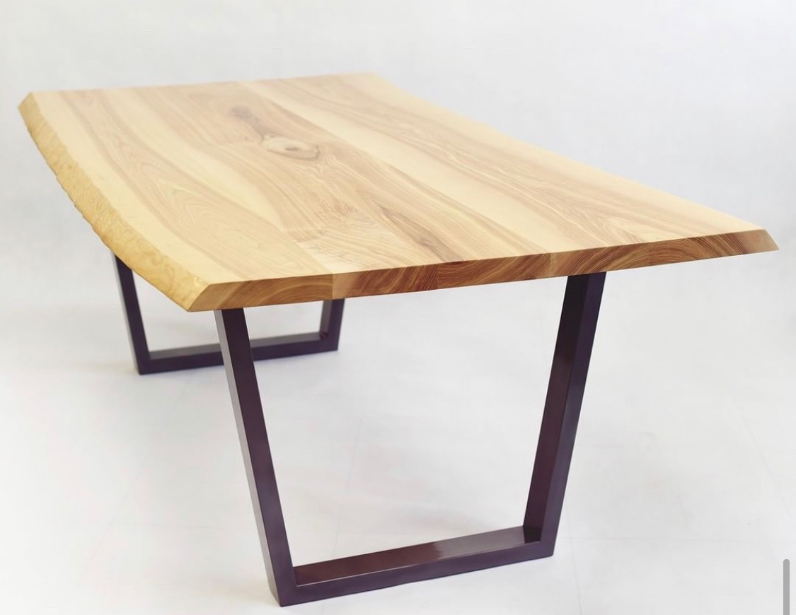 Stół z blatem z jasnego drewna o nieregularnej krawędzi, wsparty na ciemnych, metalowych nogach w kształcie litery U, na białym tle.