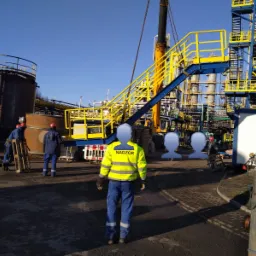 Pracownicy w kombinezonach roboczych i kaskach, w tle instalacja przemysłowa z elementami rusztowań i dźwigiem, widoczna osoba w kamizelce z napisem 'Nadzór'.