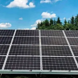 Panel słoneczny ustawiony na trawniku przed domem, widok na pola uprawne i las w tle, błękitne niebo z chmurami.