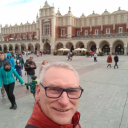 Selfie mężczyzny w okularach i czerwonej kurtce na tle Sukiennic w Krakowie, z widocznymi przechodniami.