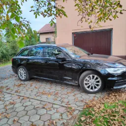 Czarny samochód kombi marki Audi zaparkowany na kostce brukowej pokrytej opadłymi liśćmi, na tle beżowego budynku z brązową bramą garażową.