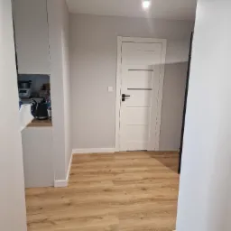 Jasne panele podłogowe w korytarzu, widoczna biała ściana, białe drzwi z czarną klamką i czarnymi poziomymi paskami, fragment kuchni w tle.