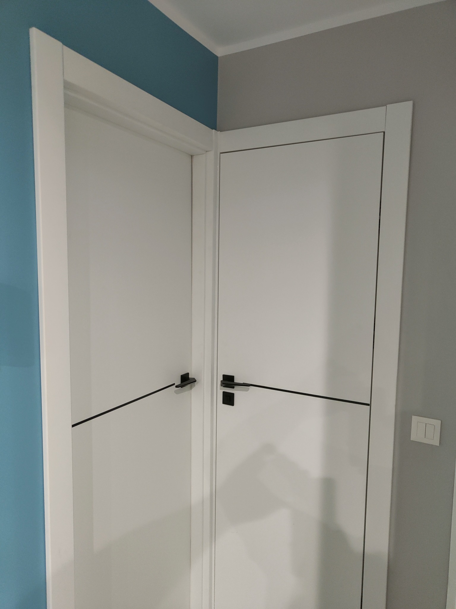 Dwa białe, nowoczesne skrzydła drzwiowe z czarnymi klamkami i poziomymi listwami, osadzone w białych ościeżnicach, na tle niebieskiej i szarej ściany.