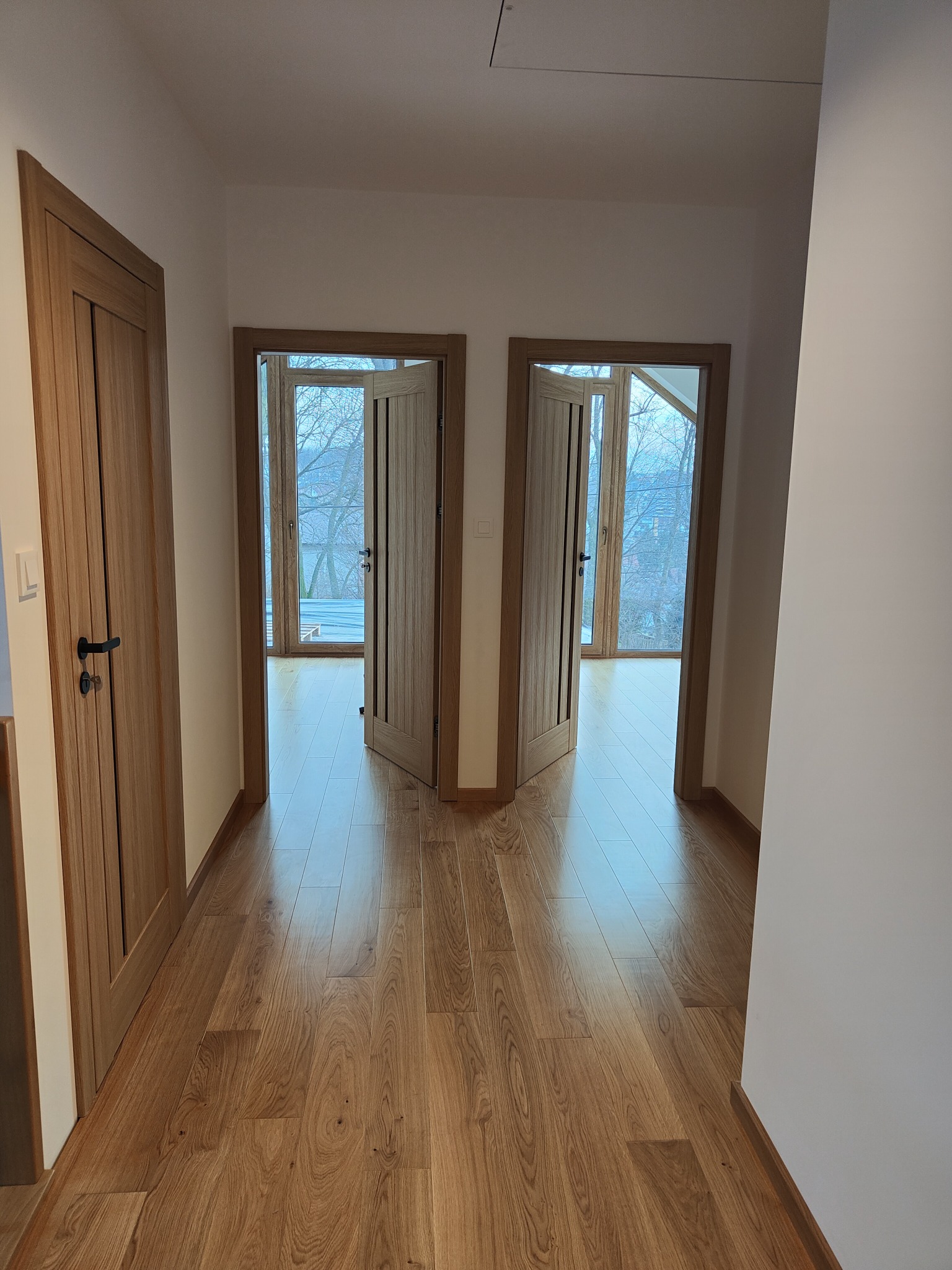 Korytarz z drewnianą podłogą i dwoma parami otwartych drzwi wewnętrznych z drewnianymi ościeżnicami, widok na zewnątrz przez okna.