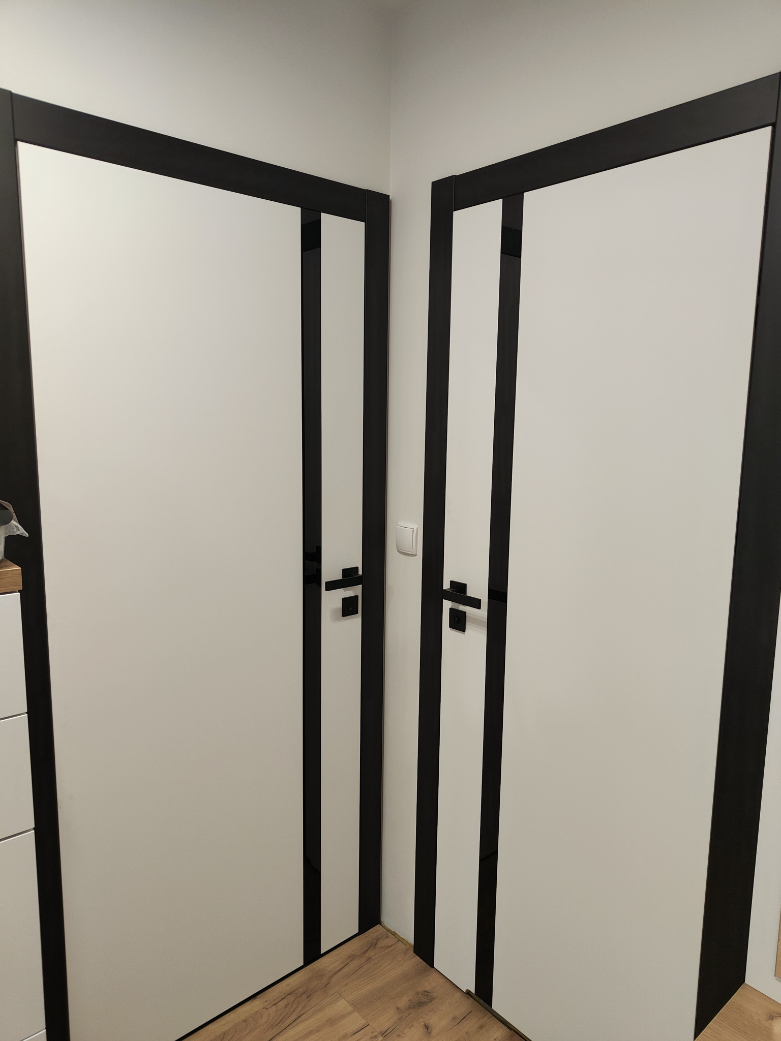 Dwie białe, nowoczesne drzwi wewnętrzne z czarnymi ramami i klamkami, ustawione pod kątem w rogu pomieszczenia, widoczny fragment drewnianej podłogi.