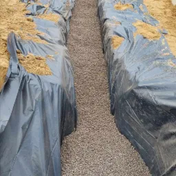 Wykop z podsypką żwirową wyłożony folią, widoczny pracownik z łopatą i niwelator laserowy na statywie.