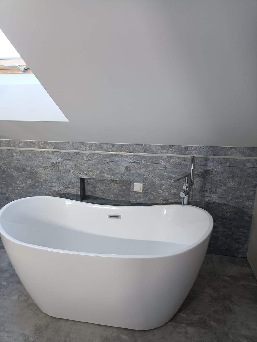 Wolnostojąca, biała wanna w łazience na poddaszu z oknem dachowym i szarą glazurą.