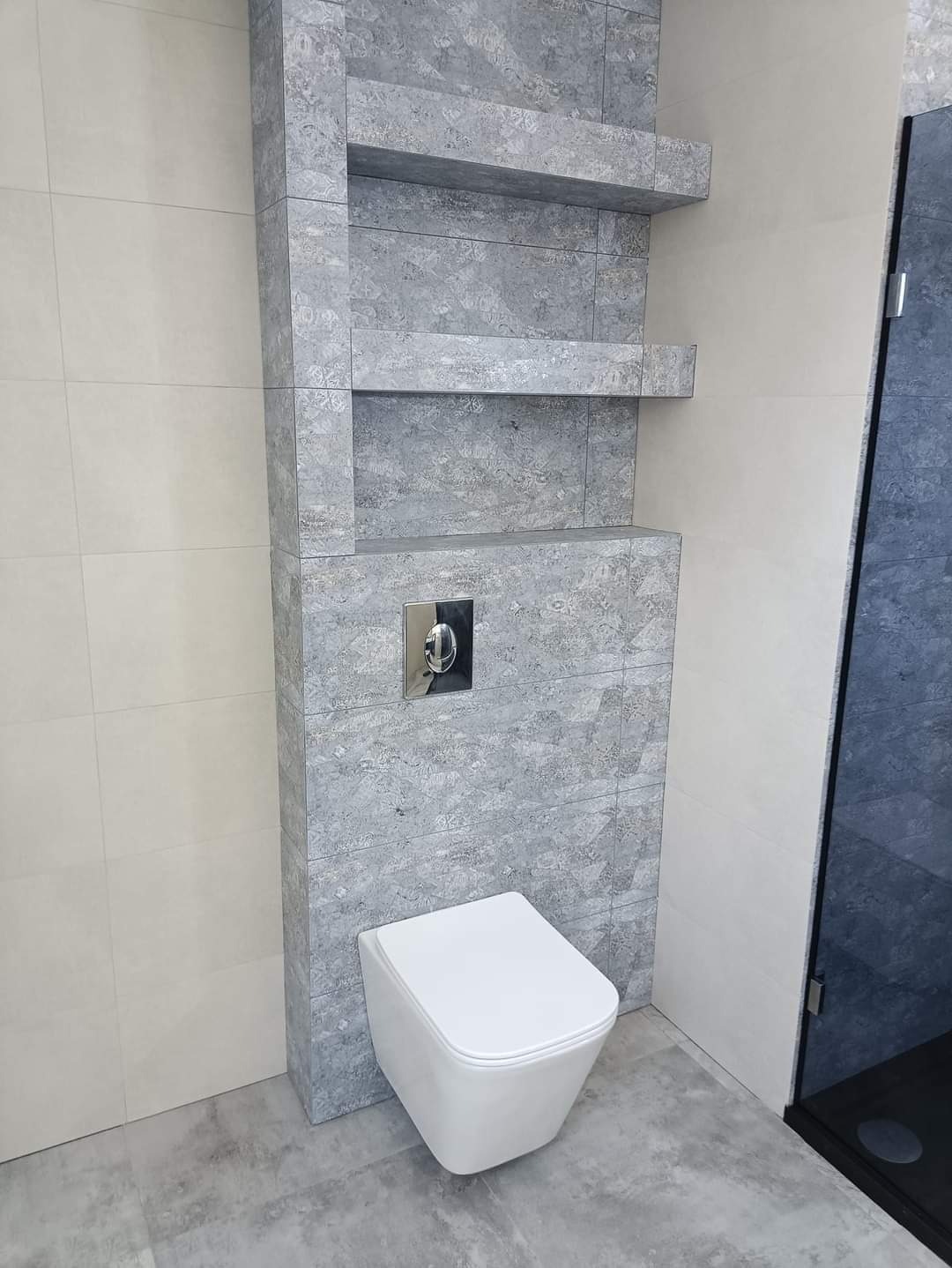 Nowoczesna łazienka z podwieszaną miską WC, szare płytki imitujące kamień na ścianie z wnękami i półkami, chromowany przycisk spłuczki.